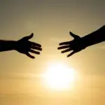 Silhouetted hands reaching towards each other with the sun setting in the background.